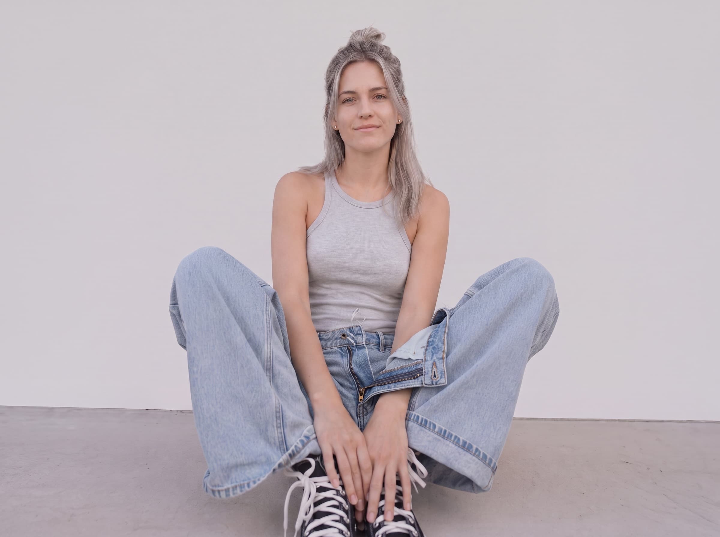 A model in a grey ribbed tank and light wash wide-leg jeans, seated against a plain studio wall